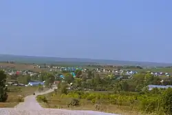 An image of a village in the distance, having buildings no larger than one story in a barely wooded region.