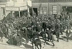 Several dozen men stand outside a plumber's shop and another building, some talking to each other and some on horseback