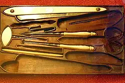 Photo of a very old waxed wood barber's kit with a dozen recessed compartments, in which some of the original tools are still to be found, including a razor, an awl and a needle. The scissors, knife and second razor are missing, thanks to the shapes in the wood. The blades are made of steel and the handles of horn or ivory.