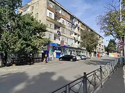 A five-story, Soviet-era apartment building with a weathered facade and numerous balconies, some with laundry hanging out to dry. The ground floor houses shops with signs. Cars are parked in front, and people are walking on the sidewalk. Tall coniferous trees stand on both sides of the building.