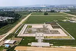An aerial view of an archaeological site amidst grassy plains near a town. In the distance, a city with high-rise apartments and a power plant looms.