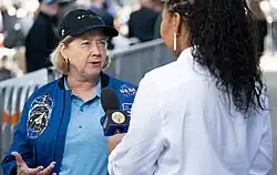 A photo of a woman with her back to the camera, wearing a white shirt and holding a microphone, interviewing a woman wearing a light blue polo shirt and a blue jacket with NASA emblem and space shuttle mission patches on it, and a black baseball hat with images representing different phases of a total solar eclipse