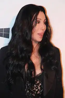 Cher, in a black lace top and blazer, with long, wavy dark hair, looks to the side with her tongue slightly out.