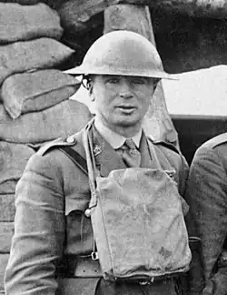 A clean-shaven man, around 40 years old but with a young-looking face. He is wearing a World War I uniform, with Brodie helmet and respirator pack. In the background is a shelter constructed out of sandbags.