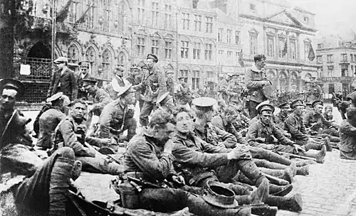In the foreground soldiers are sitting down with civilians walking behind them. In the distance are four storey buildings
