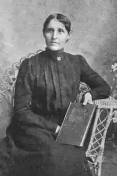 A formal portrait of an indigenous (Dakota) woman, wearing a dark dress with a high collar and long sleeves, seated in a wicker chair and holding a book