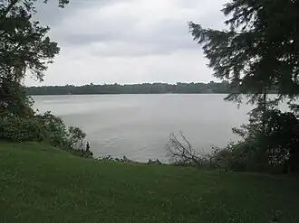 A view of a lake through a break in the trees on the near shore. The far shore of the lake is wooded.
