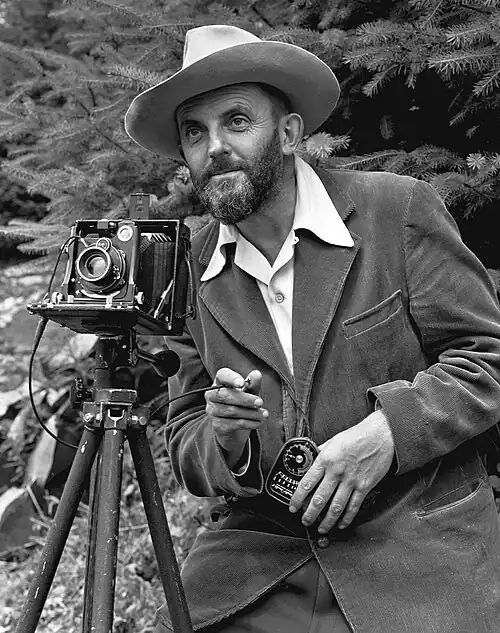 A photo of a bearded Ansel Adams with a camera on a tripod and a light meter in his hand. Adams is wearing a dark jacket and a white shirt, and the open shirt collar is spread over the lapel of his jacket. He is holding a cable release for the camera, and there is a rocky hillside behind him. The photo was taken by J. Malcolm Greany and first appeared in the 1950 Yosemite Field School Yearbook.