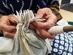 A portion of white fabric with colour-gradient blocks of woven blue pinstripes being tied into cones in the kumo shibori (spider shibori) style by a craftsperson.