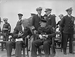 Black and white photograph of a group of officers in uniforms and white or black caps standing behind two men seated on chairs talking to each other; the one on the left is in suit and tie, the one on the right is in a naval jacket and a black cap