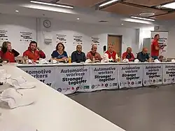 Nine trade union leaders are sitting around a table. Some wearing headsets. The table and background is adorned with English signage "Automotive workers stronger together" with logos of different union federations underneath.