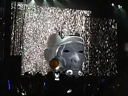 Bergling in a large prosthetic head at a stage, with a visual lights show behind him.