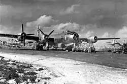 Black and white photo of a four engined propeller aircraft sitting on tarmac. Scaffolding has been erected near the nose of the aircraft and men are working on equipment nearby. The tail of another aircraft is visible to the right of the photo and buildings are visible behind the main aircraft.