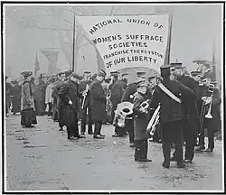 Band member assemble, reader to lead the larch. A large banner reads "National Union of Women's Suffrage Societies. Franchise the Keystone of our Liberty".