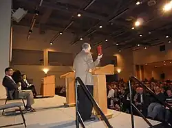 Man in his fifties standing and speaking wearing a tan suitcoat holding up a red book, man in his forties seated, large audience visible at right