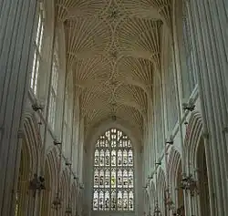 Interior of large building with a stained glass window at the far end. Above is a fan vault ceiling and on either side rows of arches.