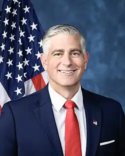 Official House portrait of Baumgartner smiling in front of the U.S. flag, wearing a black suit with American flag lapel pin, white shirt, and red tie.