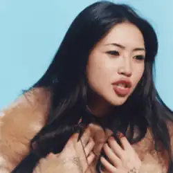 Beabadoobee, looking emphatic, mouth open, hands to chest, wearing a fur coat against a baby blue background.