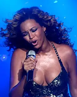 Beyoncé performing on stage, wearing a shimmering black halter dress with a plunging neckline. Her curly hair flows around her as she sings passionately into a microphone, with an intense expression on her face.