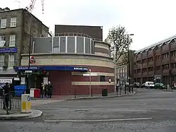 A brown- and tan-tiled building with a blue sign reading "BOROUGH STATION" in white letters and several people walking around all under a blue sky