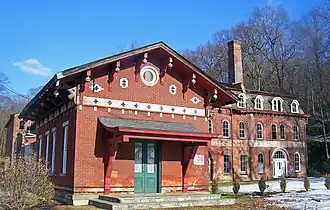 A set of brick buildings seen from slightly to their right, with some snow on the ground nearby. In the front is a small one with a pointed roof, round window and green doors. Behind is a larger one with rounded windows, a black roof and a tall chimney.