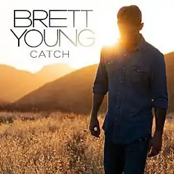 An image of a man wearing a buttoned-up denim shirt outside in a field, with the sun setting down behind him.