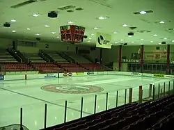 The image shows an empty Walter Brown Arena as viewed from the entrance to the arena behind the home net. The ice surface is empty. The image has a blue hue.