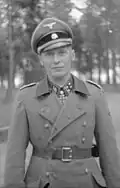 A man wearing a military uniform and coat, peaked cap and a neck order in the shape of a cross. His cap has an emblem in shape of a human skull and crossed bones.