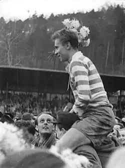 A young man in a striped shirt, smiling and holding flowers, sits on the shoulders of another young man. Both are facing away from the camera.