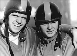 Two men wearing helmets embrace each other. The man on the left has his left arm over his partner's shoulder and grins while protecting his eyes from the direct sunlight with his brows.