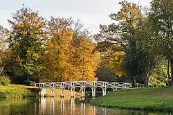 A colour photograph of a white wooden bridge across a narrow channel surrounded by trees with orange leaves