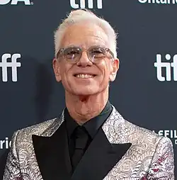 Chris Sanders, a man with short white hair and glasses and dressed in a silver and black suit and tie, smiles at the camera in front of a black sponsor board.