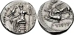 A color photograph of a silver coin on a white background. The front shows a Greek god sitting on a chair with a bird on his right hand, the back shows two horned animals