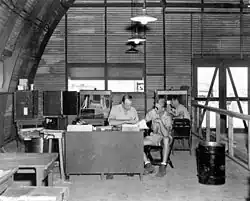 An office with desksm chairs and boxes inside a semi-cylindrical corrugated iron building.