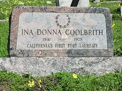 A color photograph of a pink granite headstone amid lush green grass and yellow oxalis flowers. The headstone is engraved with a laurel wreath at the top, and reads "Ina Donna Coolbrith" 1841–1928 "California's First Poet Laureate".