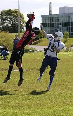 Two football players, an offensive receiver and a defensive cornerback, both reach for a thrown football. The cornerback is in front of the receiver with the ball almost secured in his hands.