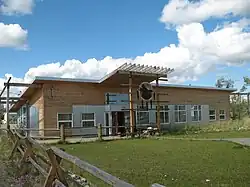 The image depicts a one-story building with a combination of wooden and corrugated metal siding with large windows on the front and a distinctive overhanging roof above the entrance, where a circular emblem is mounted. A grassy area is in front of the building. The sky is bright with a few clouds.