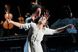 A man in dark clothes stands behind a female dancer in white