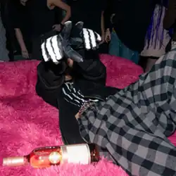 A person in pajamas lays on a pink mattress holding up their hands in skeleton gloves. A bottle sits on the mattress