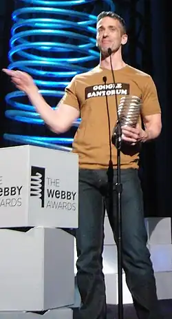 Dan Savage at the Webby Awards, holding his award and wearing a t-shirt that reads "GOOGLE SANTORUM".