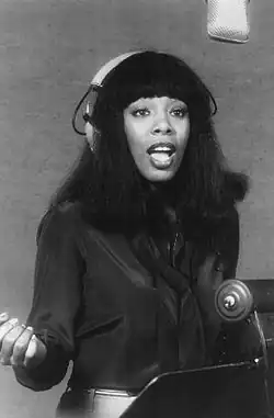 Donna Summer in a recording studio, wearing large over-ear headphones and singing into a suspended microphone. She is dressed in a dark satin blouse with a matching scarf, and her straight, shoulder-length hair features blunt bangs. Her expressive posture suggests she is mid-performance.