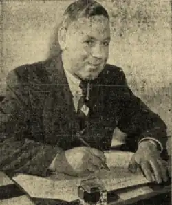 Black and white photo of Grimston in a suit and tie, sitting at a desk with pen and paper