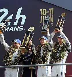 Colour photograph of four men in racing suits holding aloft trophies, with large leaf garland hung around their necks.