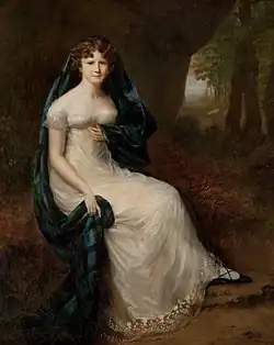 Full length portrait of young woman wearing a white empire-waist dress with a plaid of tartan loosely draped over her head and left shoulder, seated in a park