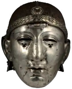 Colour photograph of the Emesa helmet, which has a silver face mask shaped like a human face, and an iron headpiece. The headpiece is decorated with a laurel-wreath diadem and a rosette, and has a central hinge holding the face mask.