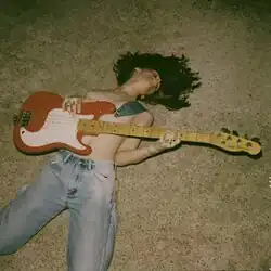A topless young woman in jeans lying down on a carpet floor holding an electric guitar