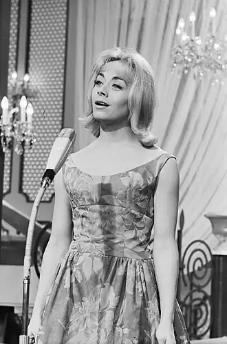 A young woman with shoulder-length blond hair and a floral dress stands in front of a microphone. There are curtains and two decorative chandeliers in the background.