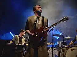 Lead vocalist, Jonathan Higgs, stands with a guitar in front of drummer, Michael Spearman, on a blue-lit stage.
