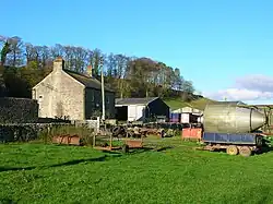 A green field with farm buildings and farm equipment, with a small hill behind
