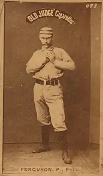 A baseball-card photograph of a mustachioed man in an old-style white baseball uniform holding a baseball in front of his chest with both hands